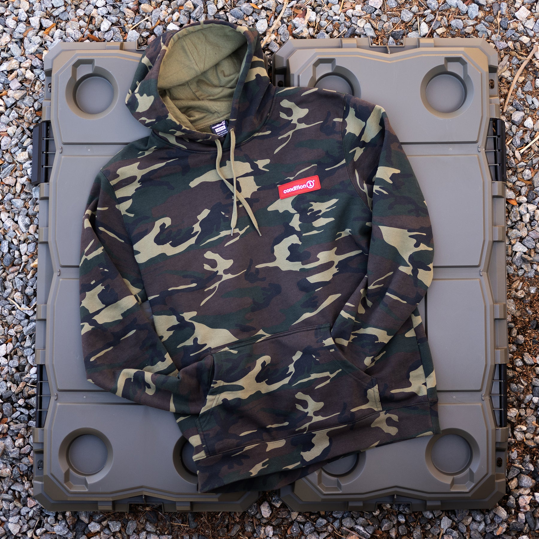 Camouflage hoodie with concealed pockets and a red "Condition" logo patch, laid on a gray plastic surface outdoors.