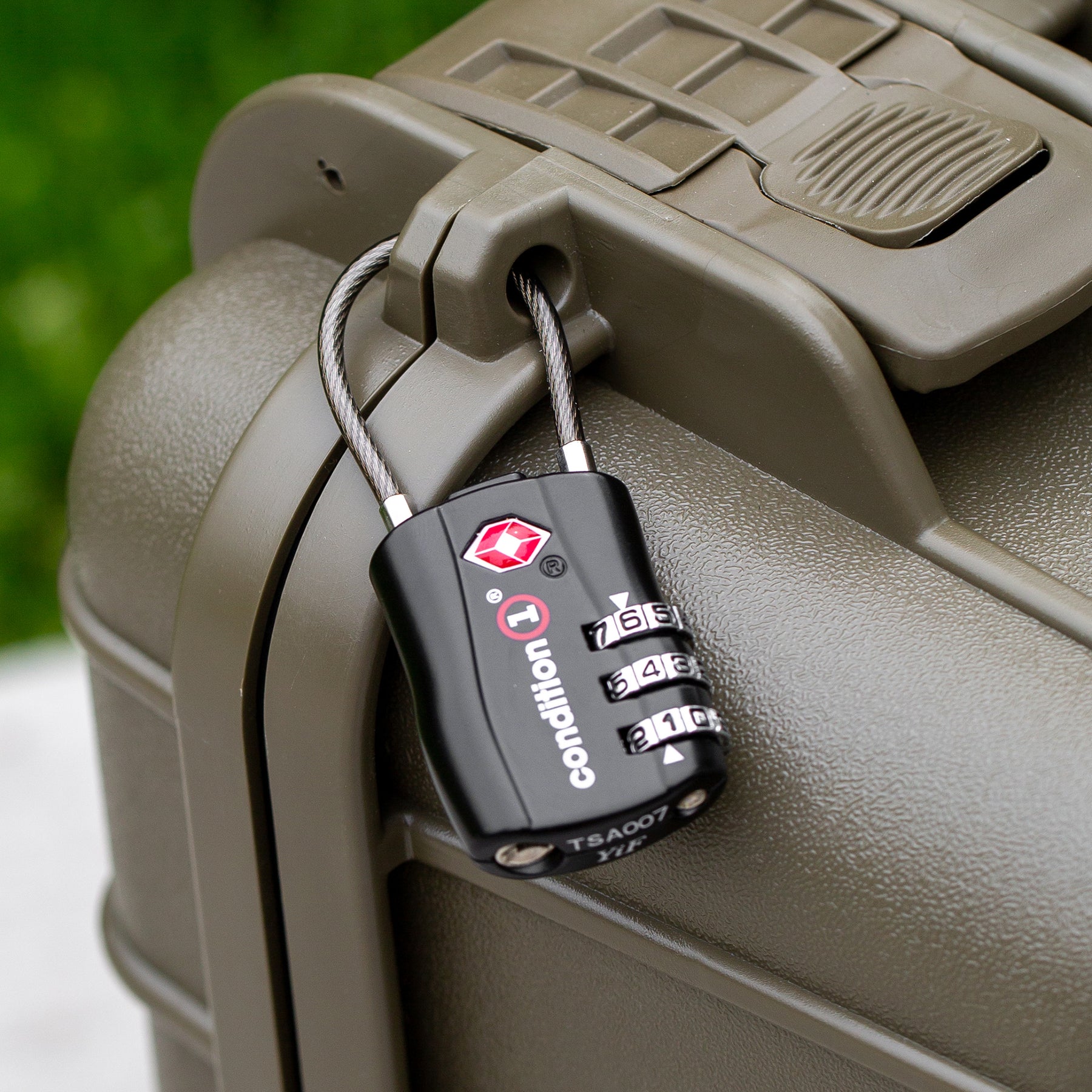Two black combination locks securing a gray hard-shell suitcase, with a green outdoor background.
