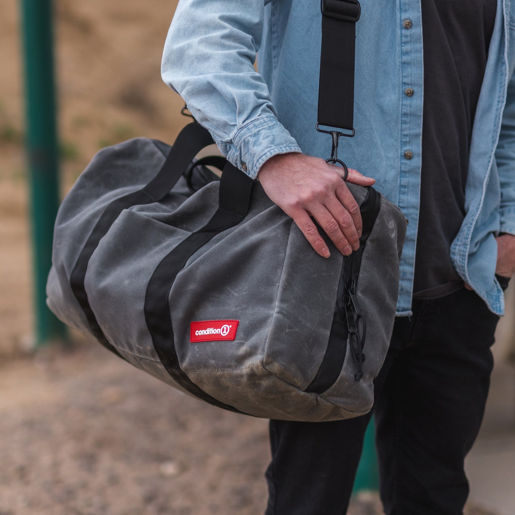 Close-up of a person holding a gray waxed canvas duffle bag with black straps and a red "condition 1" logo.