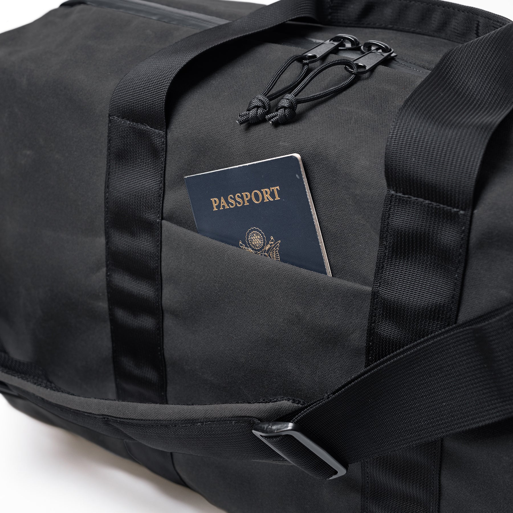 Black waxed canvas duffle bag with a front pocket holding a passport, featuring sturdy black straps and zipper details.
