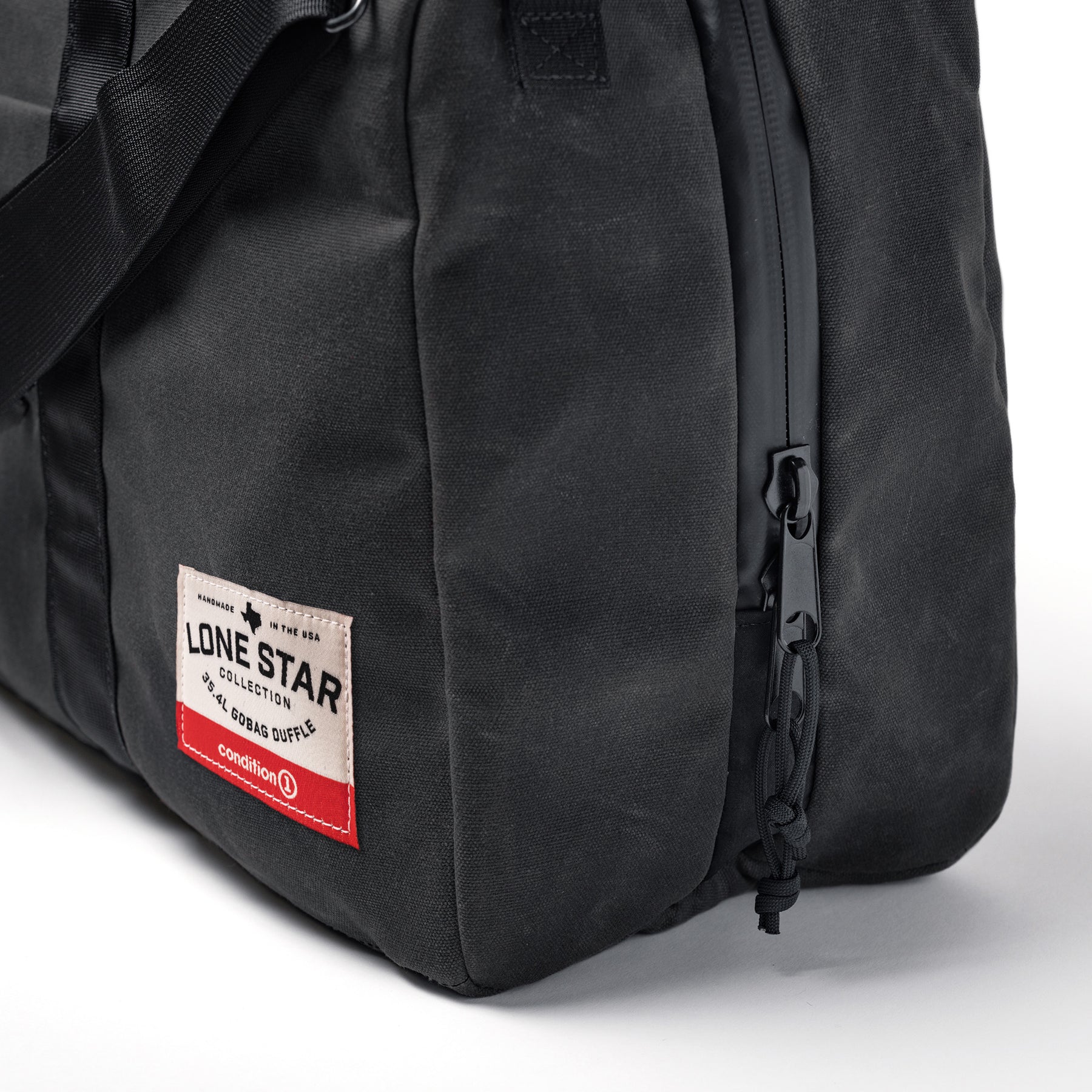 Close-up of the black waxed canvas duffle bag with a front zipper pocket and a white, red, and black label reading "Lone Star Collection.