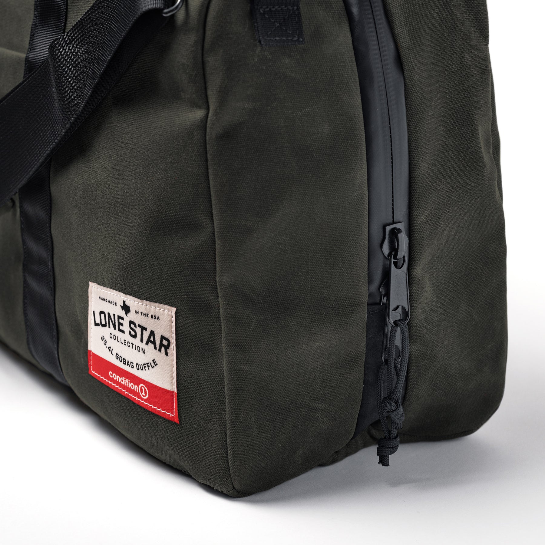Dark green waxed canvas duffle bag with black zipper, front patch label, and black strap, designed for travel and durability.