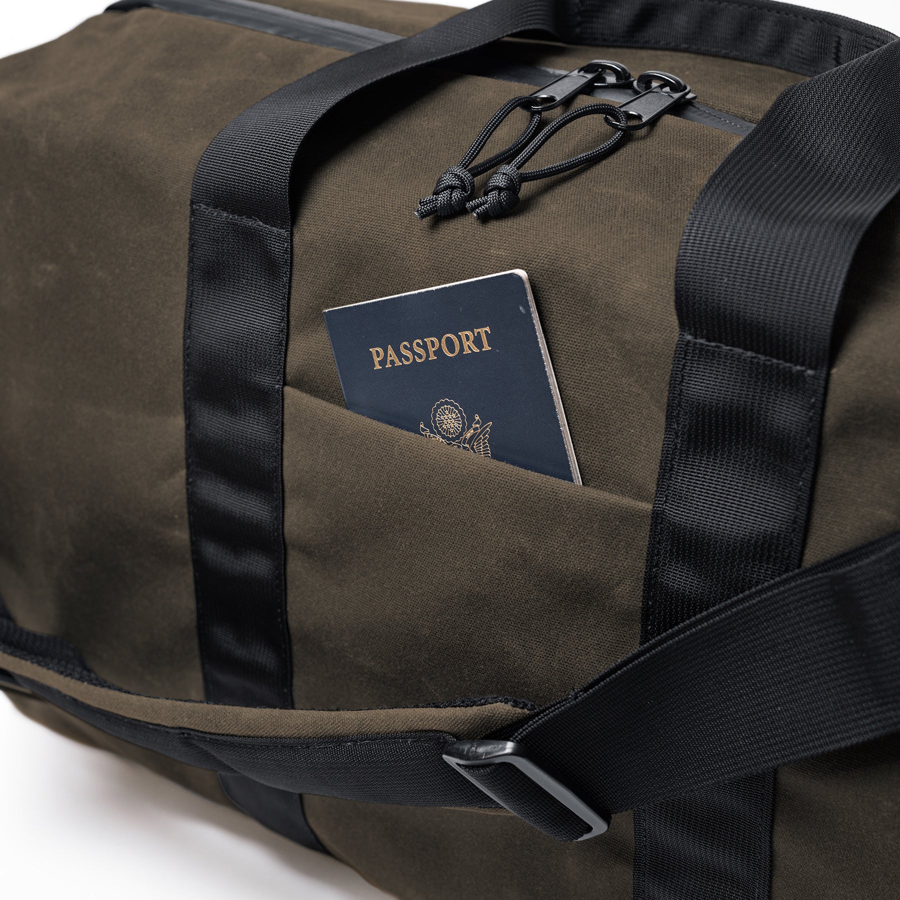 The GOBAG Waxed Canvas Duffle bag with a passport in an exterior pocket, featuring black straps and zipper pulls.