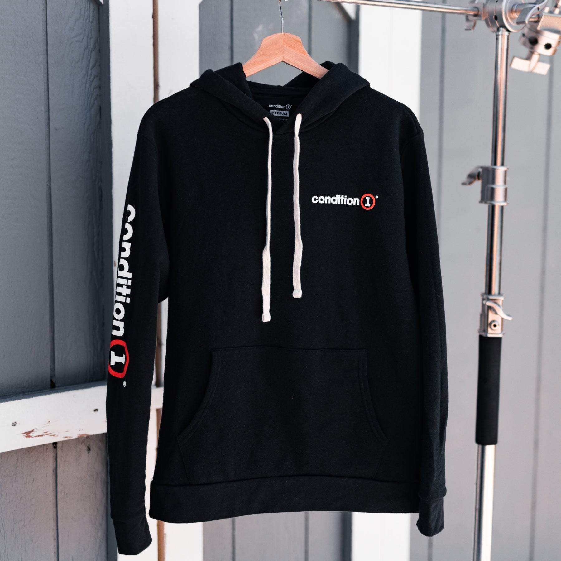 Black hoodie with "condition" logo on chest and sleeve, featuring a front pocket and white drawstrings, hanging on a wooden hanger.
