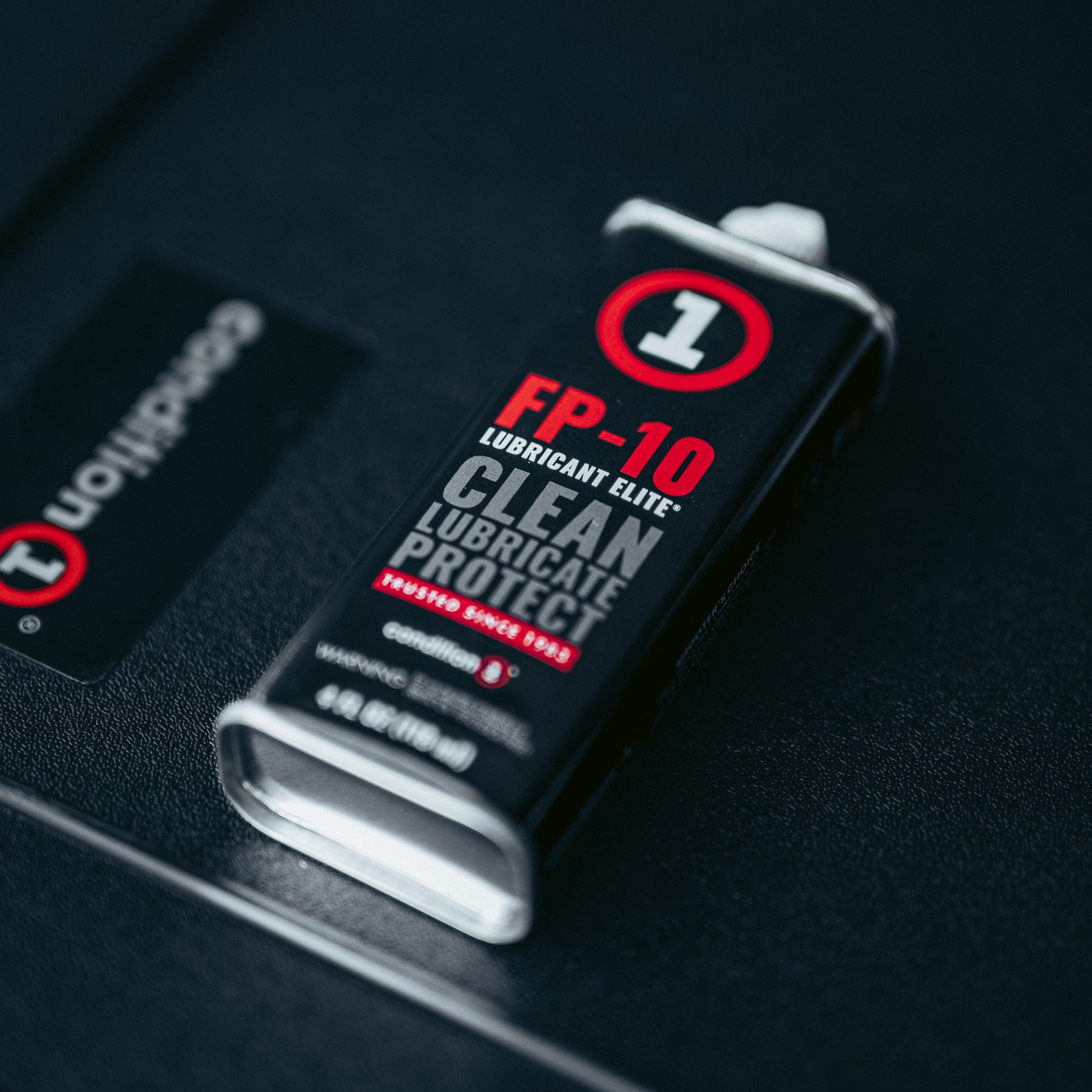 A black and red bottle of Condition 1 Elite CLP Firearm Oil with "Clean, Lubricate, Protect" labels, placed on a textured black surface.