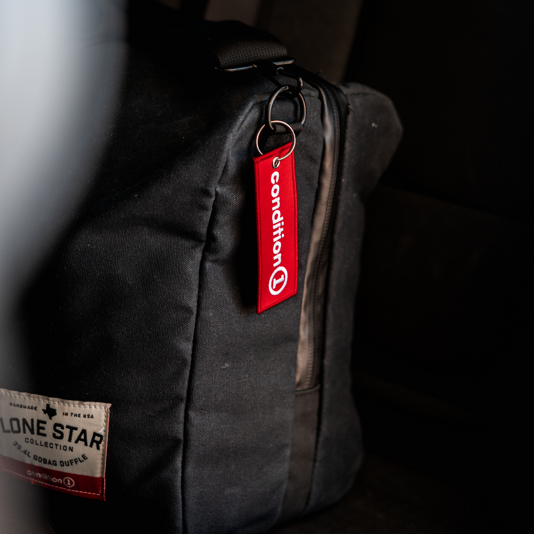 Red Condition 1 keychain attached to a black backpack, featuring a metal ring and a fabric tag with white text.