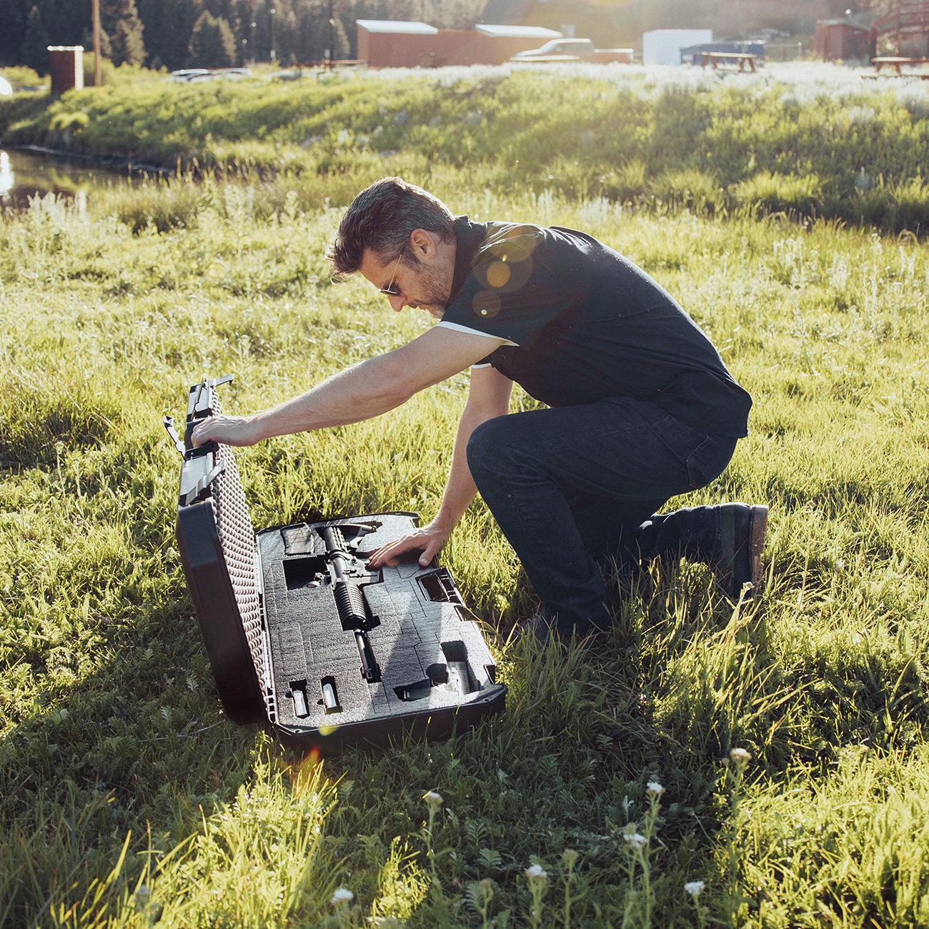 Man kneeling on grass opening a black hard case containing a firearm, outdoors in a sunny, grassy field.