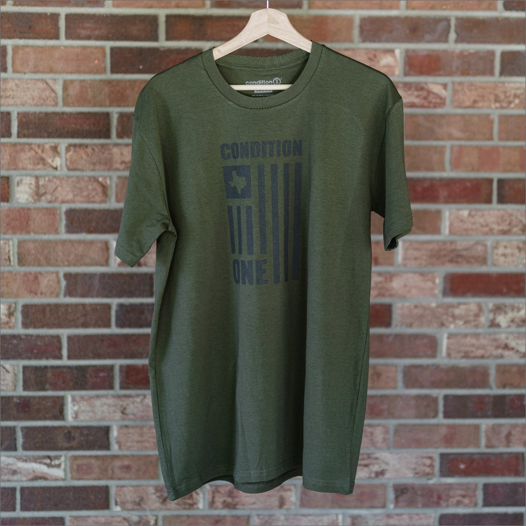 Olive green T-shirt with "Condition One" text and a flag graphic printed on the front, hanging on a wooden hanger against a brick wall.