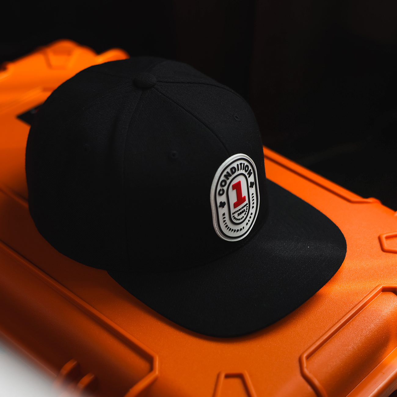 Black snapback hat with a white and red "Condition 1" logo patch, resting on an orange case against a dark background.