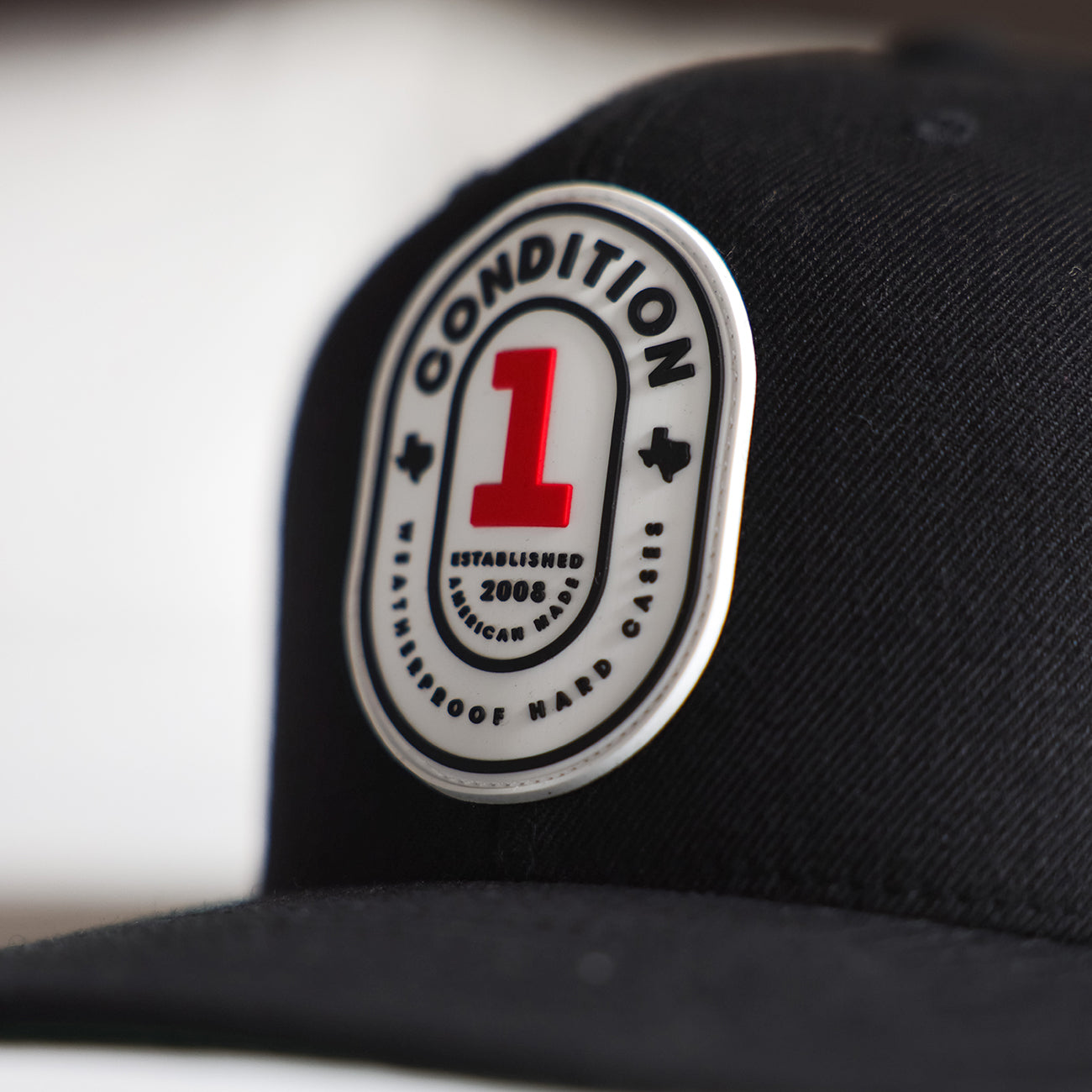 Close-up of a black snapback cap featuring a circular patch with "The One" branding, a large red number 1, and "Established 2008" text.