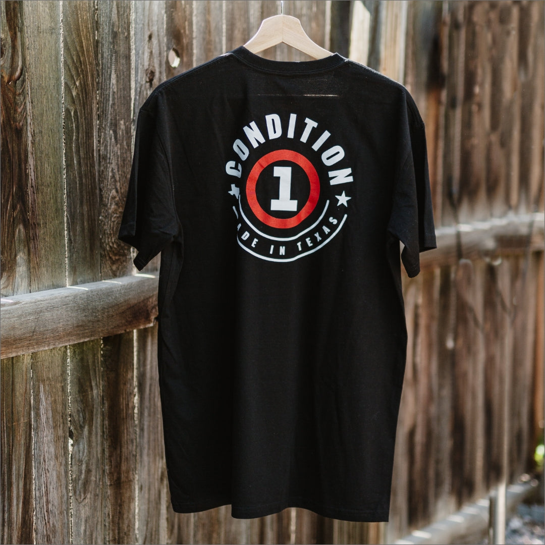 Black T-shirt with "Condition 1 Made in Texas" graphic design on the back, hanging on a wooden fence.