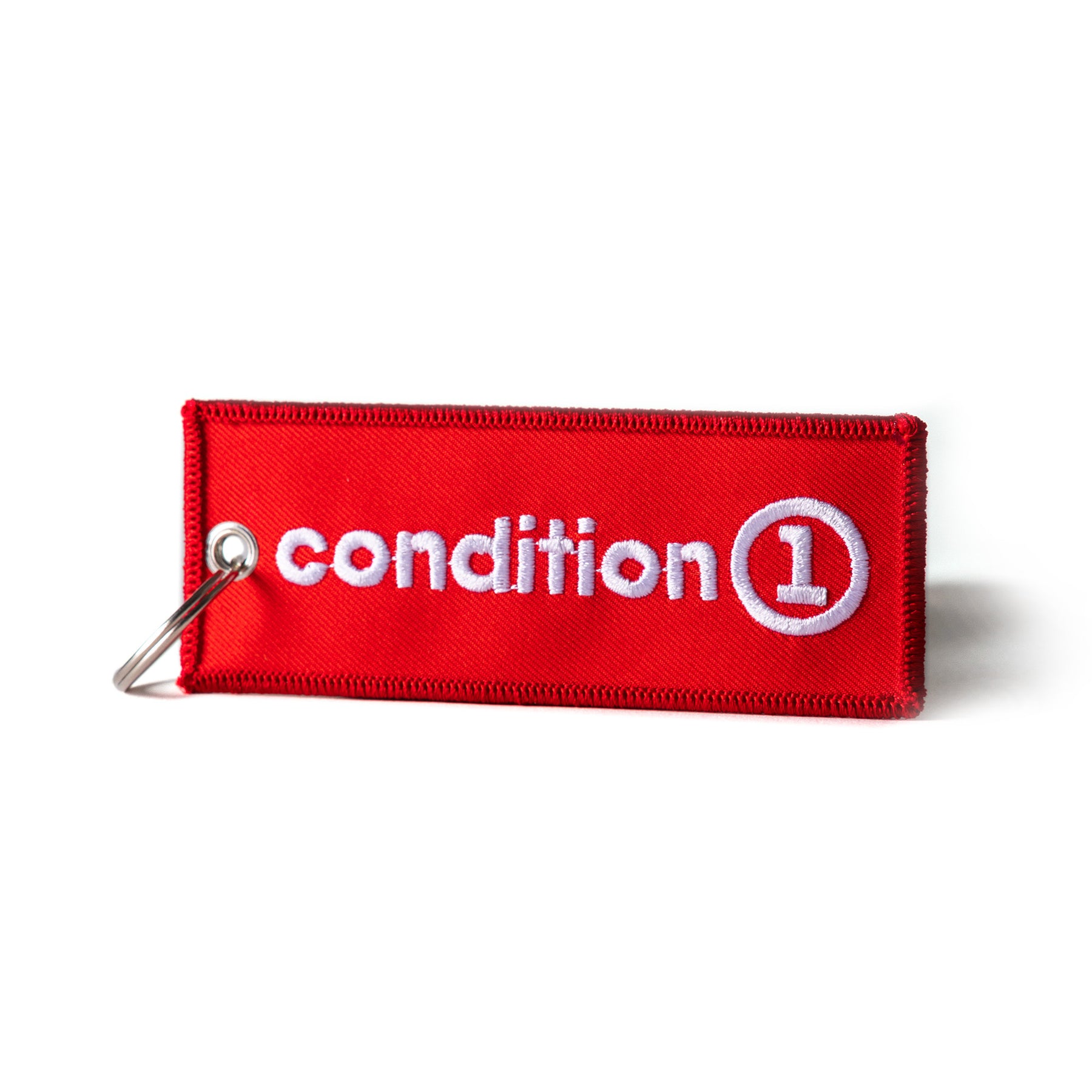 Red fabric keychain with white embroidered "condition 1" text and a circular logo, attached to a metal keyring.