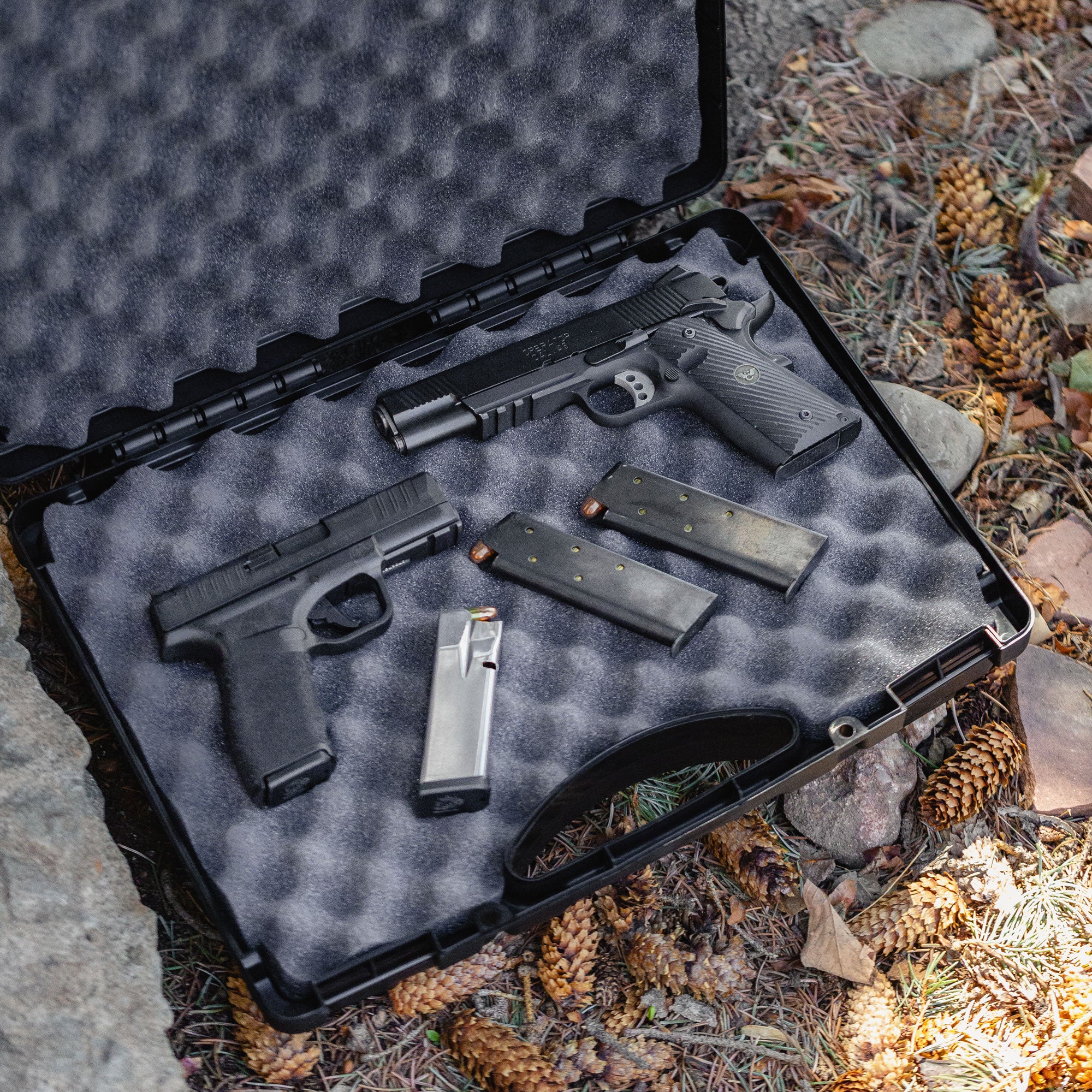 Alt text: "Open 17-inch Economy Case #1764 displaying two pistols, several magazines, and foam padding, set on a forest floor with pine cones around."