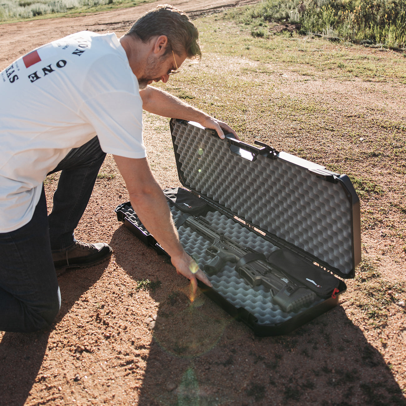 Man opening a black 42" Economy AR Case #2300 with foam padding, outdoors on dirt ground, holding a rifle inside.