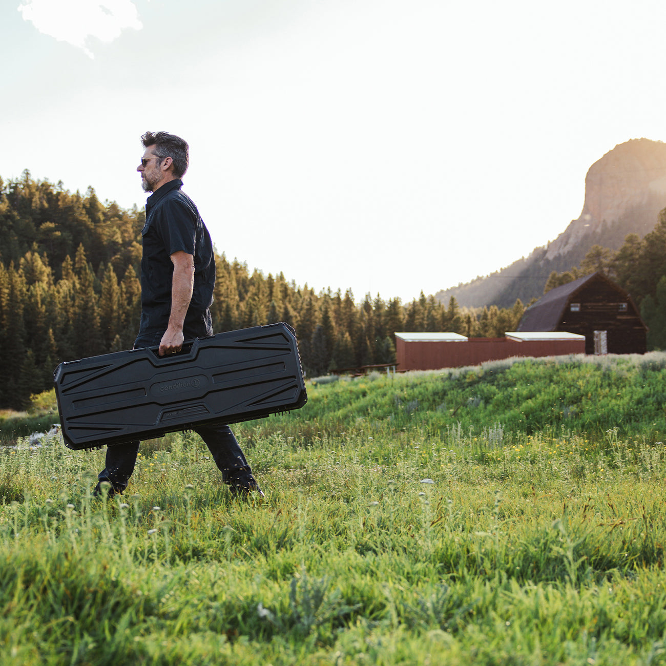 Man walking outdoors with a black 42" Economy AR Case #2300, carrying it in a scenic grassy field with mountains in the background.