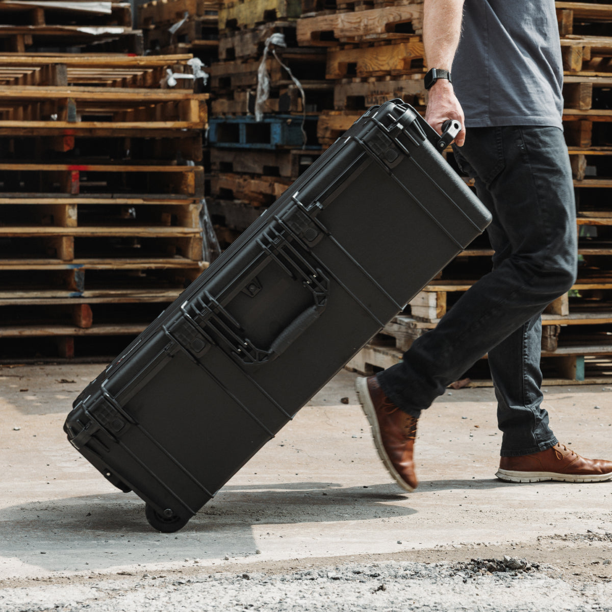 Person wheeling a black hard case across a concrete floor near stacked wooden pallets